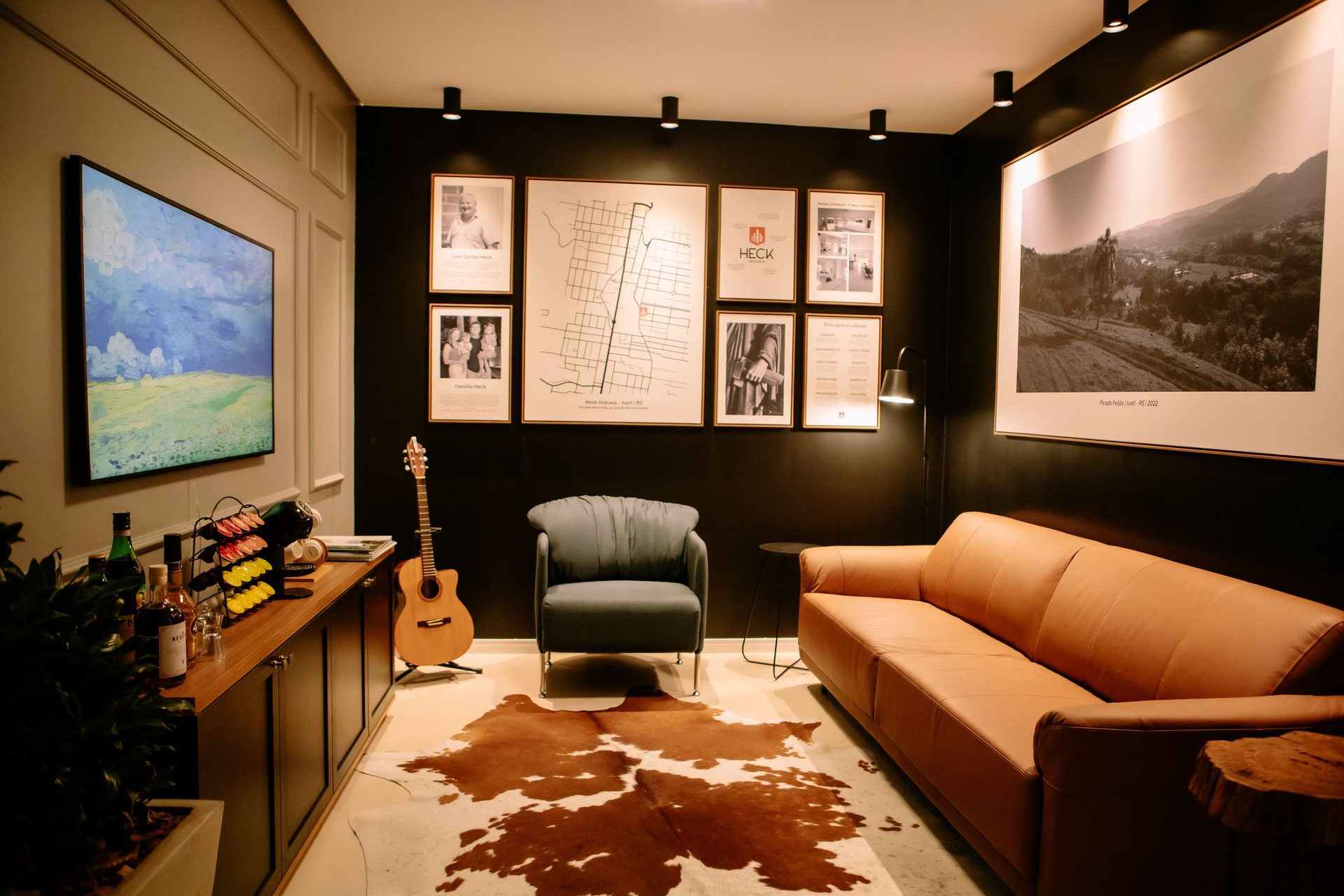 Sala de atendimento da Heck Imóveis em Ivoti: parede preta com a galeria da família e do mapa da cidade, poltrona, sofá de couro caramelo e tapete.